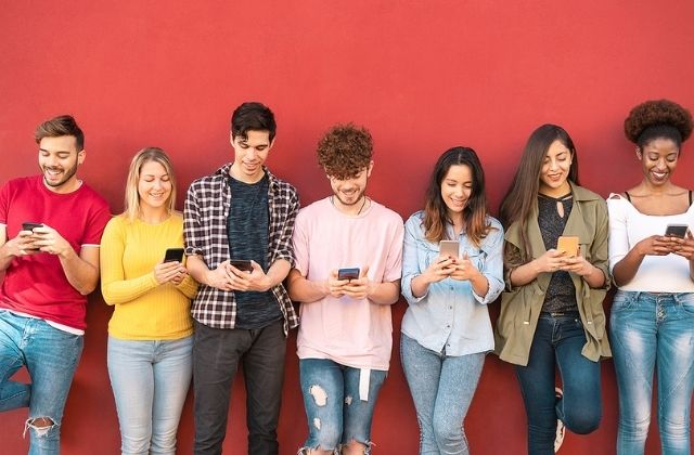 A diverse group of students leaning against a red wall, focused on their smartphone.